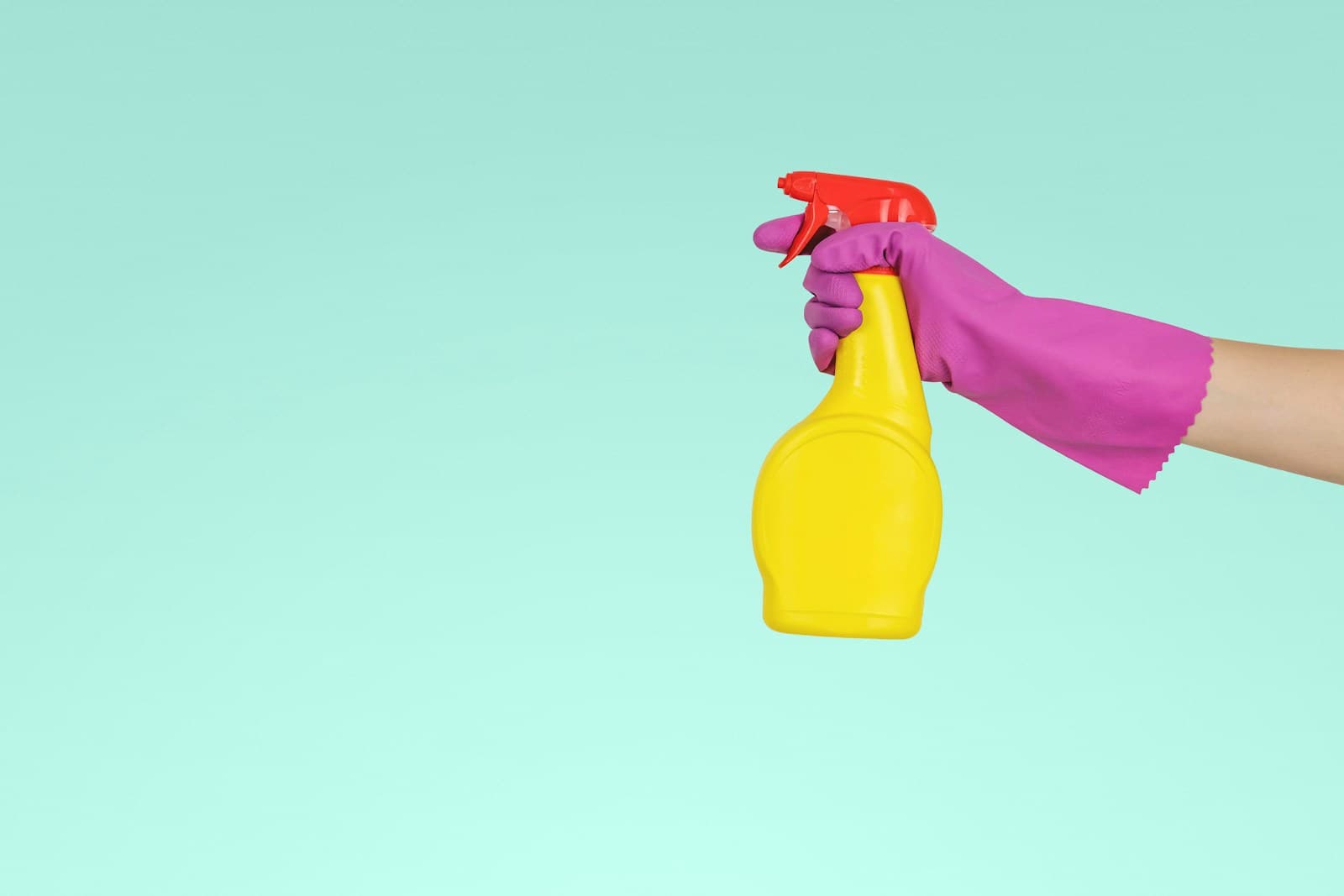 Gloved hand holding a yellow spray bottle against a mint background