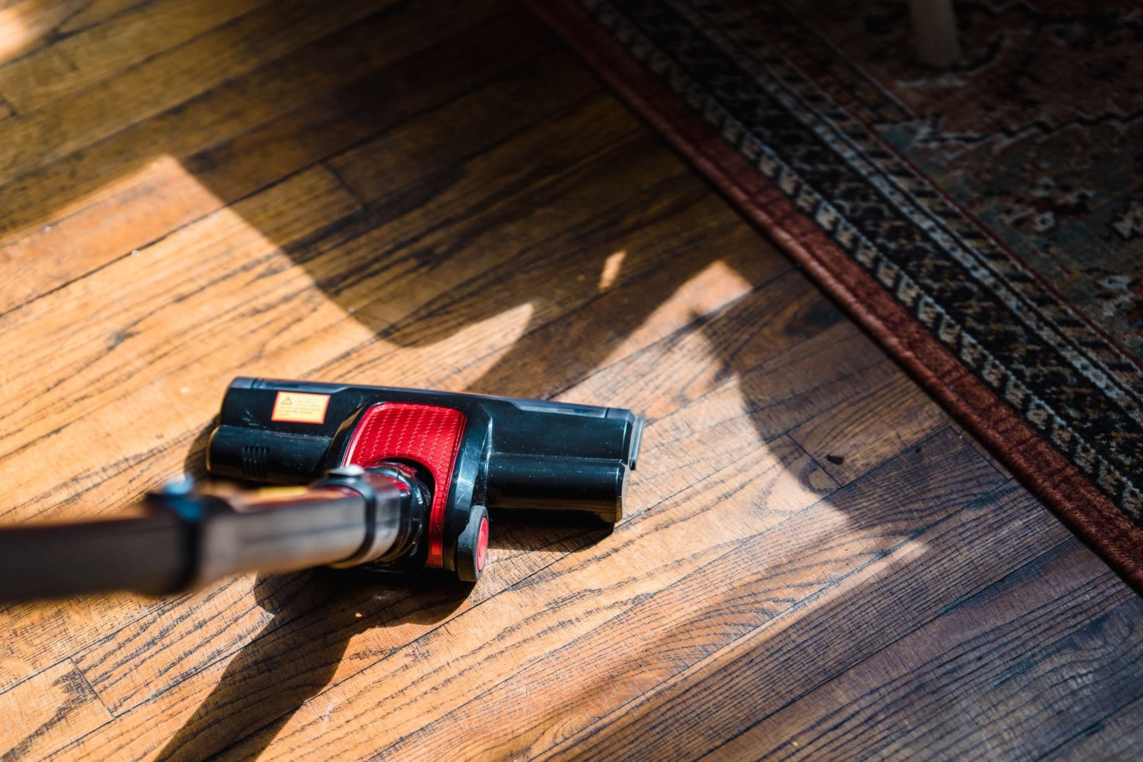 A hand pressing a vacuum head across a sunlit hardwood floor