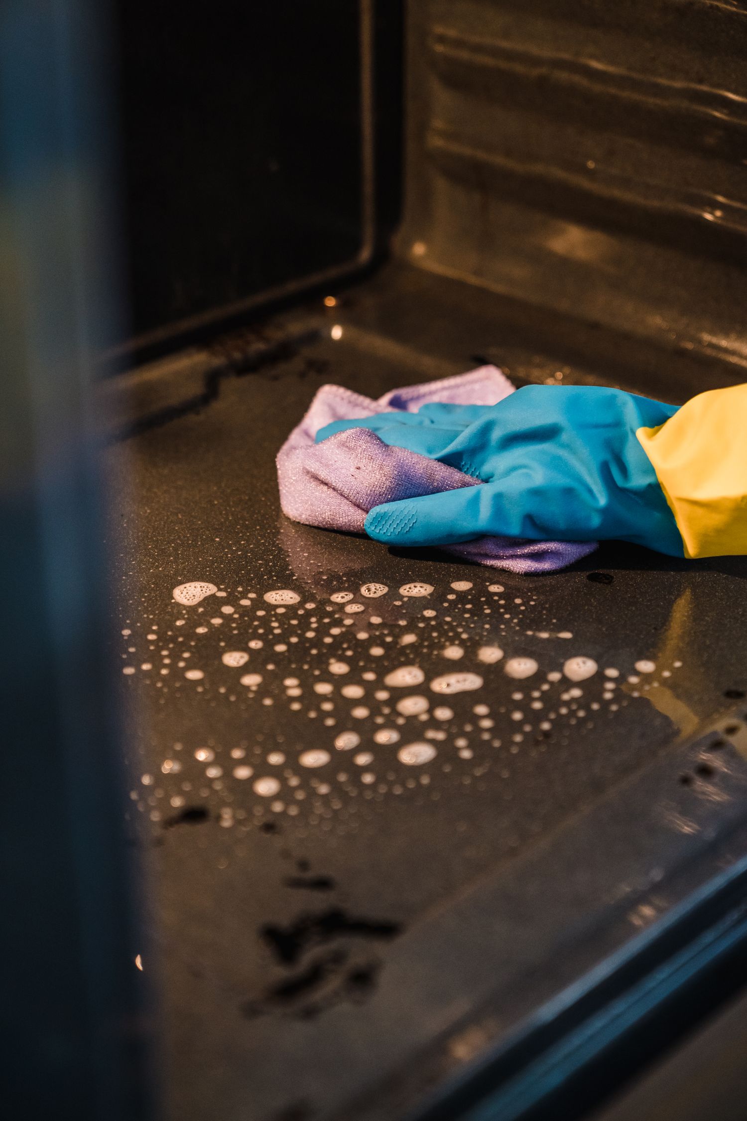A gloved hand wiping suds inside an oven with a cloth